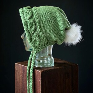 Puede incluir: Un gorro de punto color verde salvia con un patrón de hojas texturizado en el borde frontal, dos largas trenzas colgando de los lados y un pompon de piel sintética blanca esponjosa en la parte superior trasera del gorro.