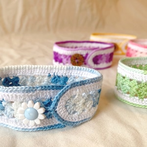 May include: A set of five crocheted bracelets with different coloured patterns and buttons. The bracelets are made with white, blue, green, purple and pink yarn.