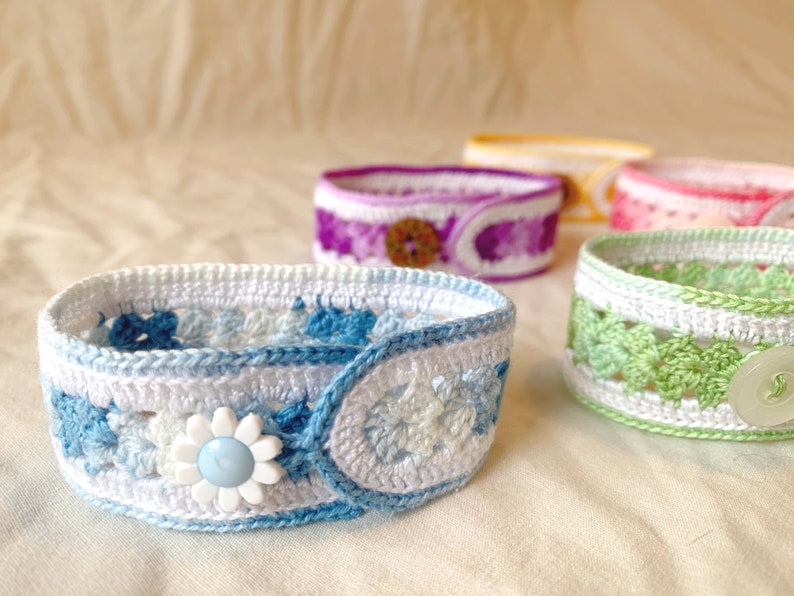 May include: A set of five crocheted bracelets with different coloured patterns and buttons. The bracelets are made with white, blue, green, purple and pink yarn.