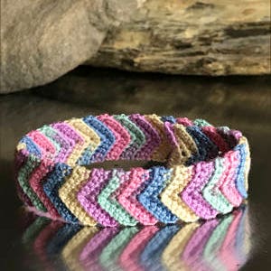 May include: A multi-colored chevron patterned crocheted bracelet with shades of pink, blue, green, and beige.