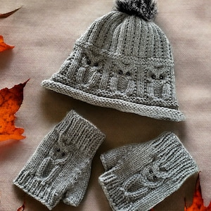 May include: A gray knitted hat with a fluffy pom pom and matching fingerless gloves. The hat and gloves have a cable knit pattern and are decorated with small black buttons. The items are displayed on a beige background with autumn leaves.
