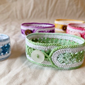 May include: Five crocheted bracelets with different coloured patterns and white buttons. The bracelets are made with a variety of stitches and colours, including blue, green, pink, purple, and yellow.