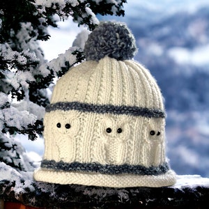 May include: White knitted hat with a gray pom pom and a gray stripe pattern. The hat has a design of three owls with black eyes.