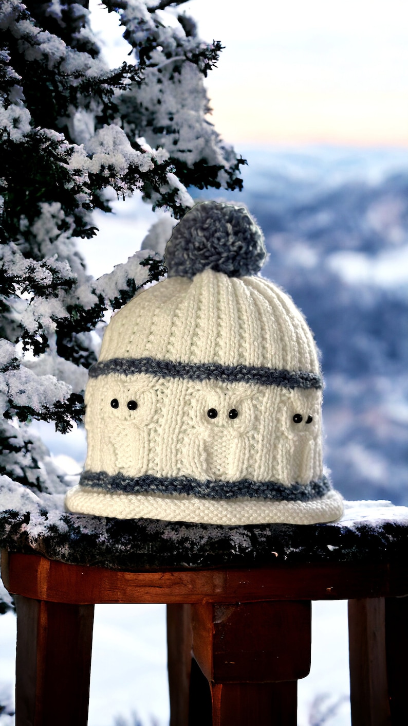 May include: White knitted hat with a gray pom pom and a gray stripe pattern. The hat has a design of three owls with black eyes.