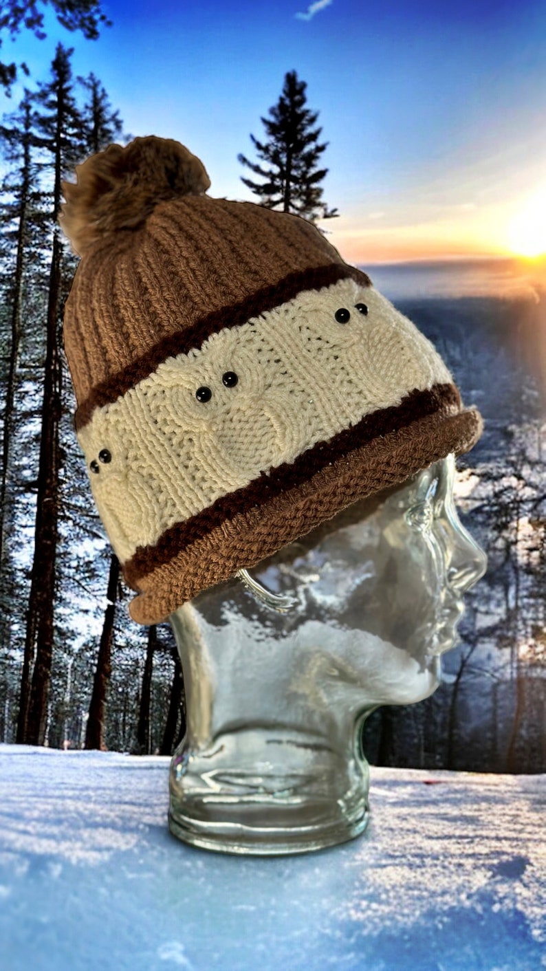 May include: A brown and white knit beanie with a fluffy pom pom on top. The beanie has a brown band with black buttons.