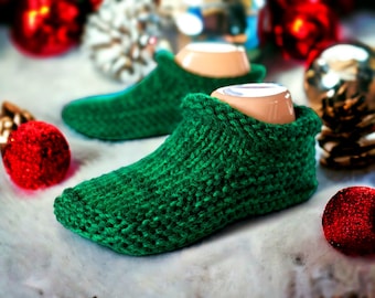 May include: A pair of green knitted slipper socks with a textured knit pattern. The slippers are on a white surface with red and gold ornaments in the background.