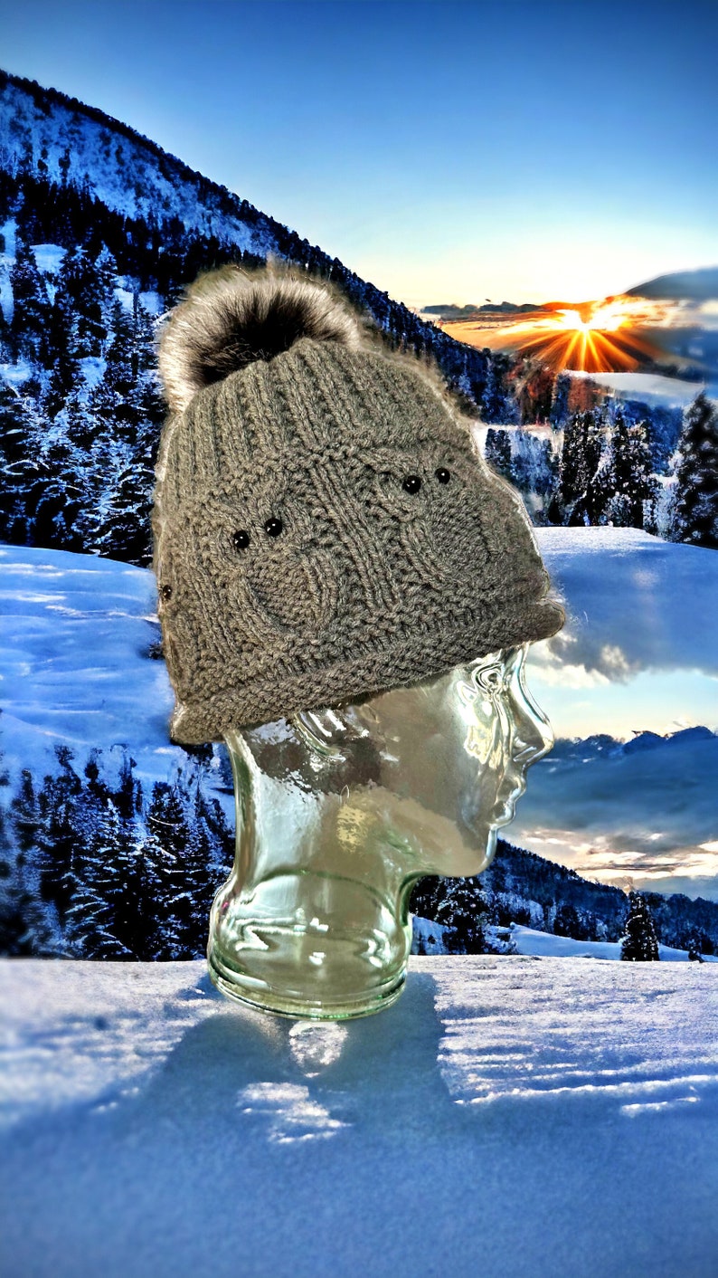 May include: A gray knitted beanie with a faux fur pom pom and black embellishments. The beanie is on a glass mannequin head against a snowy mountain backdrop.