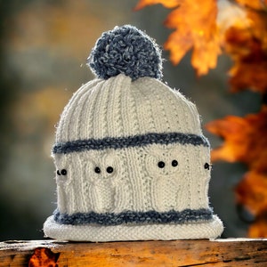 May include: White knitted hat with a blue pom pom and owl designs. The hat is sitting on a wooden surface with fall leaves.