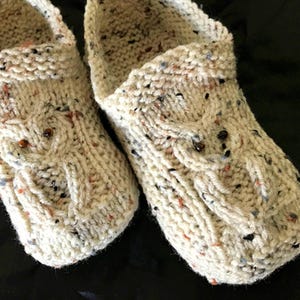 May include: A pair of knitted cream-colored slippers with a speckled pattern. The slippers have a cable knit design and are decorated with small brown beads.