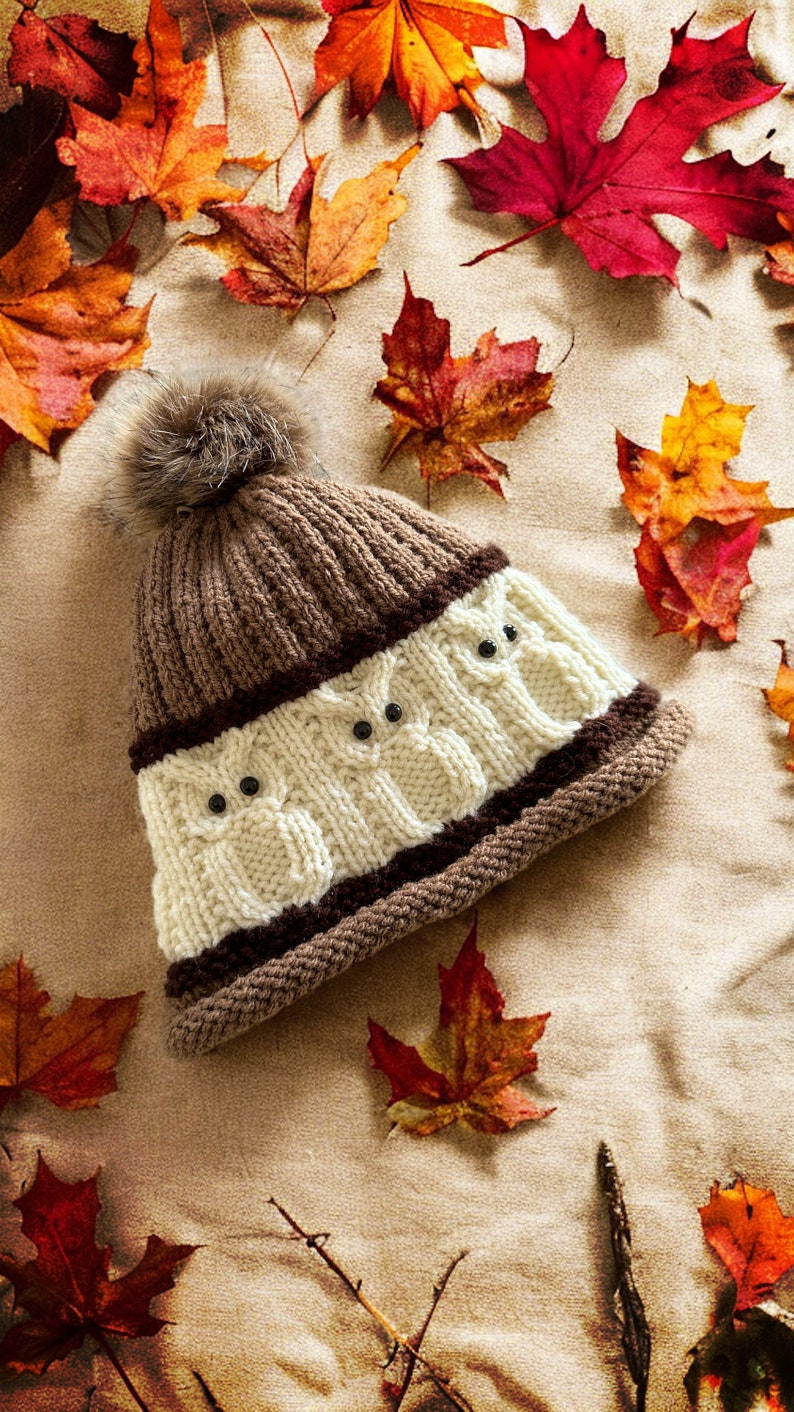 May include: A brown and white knitted hat with a fluffy pom pom on top. The hat features a pattern of owls with black eyes. The hat is lying on a beige surface with autumn leaves scattered around it.