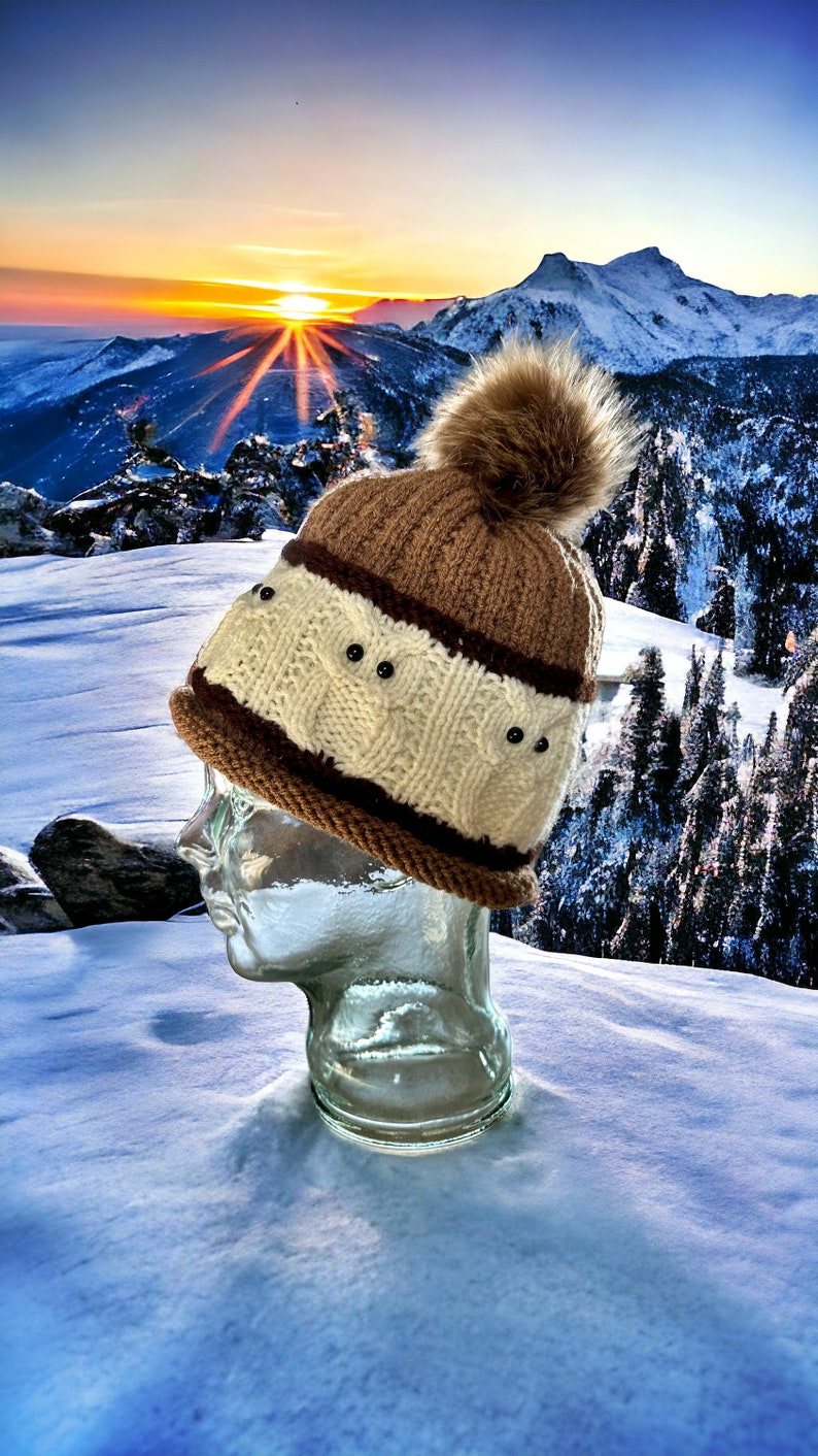 May include: A brown and white knit hat with a brown pom pom. The hat has a cable knit design and is accented with black beads. The hat is sitting on a glass head form against a snowy mountain background.