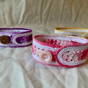 May include: Three crocheted bracelets with different coloured patterns and buttons. The bracelets are white with purple, pink, and yellow accents.
