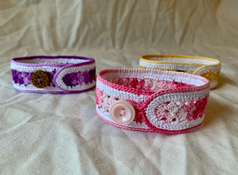 May include: Three crocheted bracelets with different coloured patterns and buttons. The bracelets are white with purple, pink, and yellow accents.