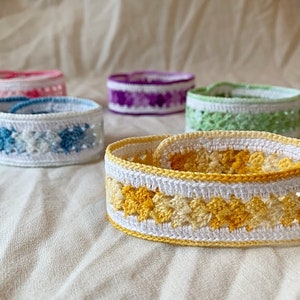 May include: A set of six crocheted bracelets in various colours, including yellow, blue, green, and purple. The bracelets are made with a white yarn and feature a variety of crochet stitches.