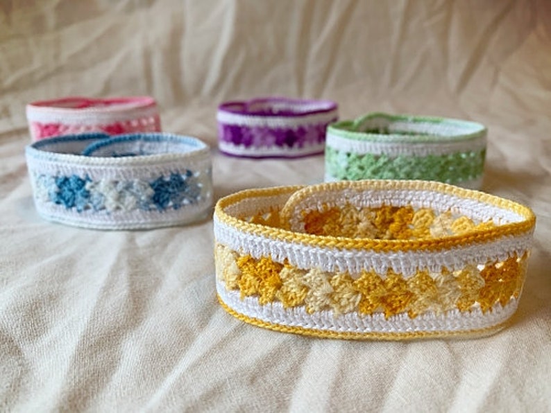 May include: A set of six crocheted bracelets in various colours, including yellow, blue, green, and purple. The bracelets are made with a white yarn and feature a variety of crochet stitches.