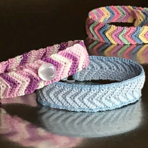 May include: Three crocheted bracelets with a chevron pattern. The bracelets are in shades of pink, blue, and multi-colored. One bracelet has a white button.