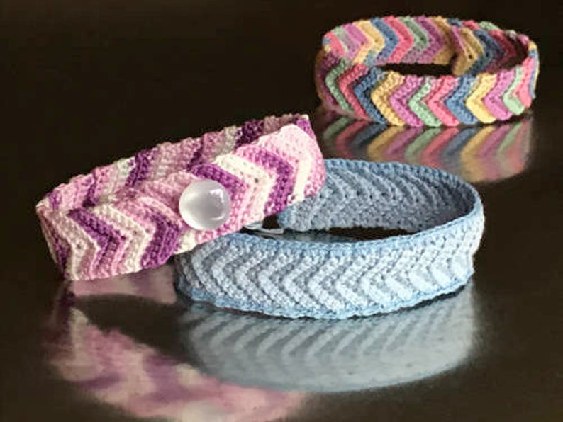 May include: Three crocheted bracelets with a chevron pattern. The bracelets are in shades of pink, blue, and multi-colored. One bracelet has a white button.