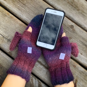 May include: A pair of purple and blue knitted fingerless gloves with a white patch on each glove. A white smartphone with a black screen is resting on top of the gloves.