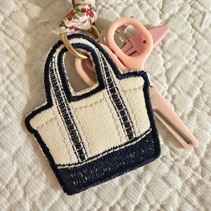 May include: A small, cream-colored tote bag keychain with navy blue trim and accents. The keychain has a gold ring and a floral fabric strap. A pair of pink scissors is also visible.