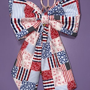 May include: A large, decorative bow with a patchwork design in red, white, and blue. The bow features patterns including stripes, floral prints, and stars, evoking a patriotic theme. It is likely used for decoration.