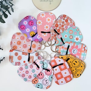 May include: A collection of colorful sunglass cases with various patterns, including smiley faces and floral designs. Each case is attached to a silver keyring. The cases feature the text "Good Vibes" and "The Accessory Design Co."