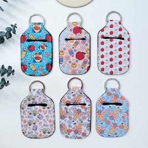 May include: Six different colourful hand sanitiser holders with keychains. Each holder features a different back-to-school themed pattern, including apples, pencils, school buses, and bows. The holders are made of a soft, flexible material and are perfect for keeping your hands clean on the go.