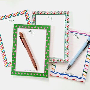 May include: Four personalised notepads with decorative borders and pens. The notepads feature initials and names, with colourful patterns including hearts, checks, and floral designs. One notepad has a brown pen, and another has a light blue pen.
