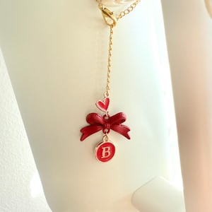May include: A gold chain with a red heart, red bow, and a red circle with the letter "B" in gold. The charm is hanging from a white surface.