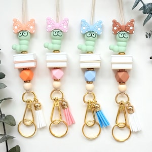 May include: Four colorful beaded lanyards with gold-tone hardware and tassels. Each lanyard features a green caterpillar charm with a bow tie, a stack of white blocks, and a geometric bead in orange, pink, blue, and brown. The tassels are white, pink, and blue.