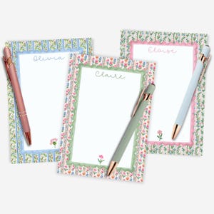 Puede incluir: Tres blocs de notas personalizados con bordes florales y bolígrafos a juego. Los blocs de notas tienen los nombres de Olivia, Claire y Eloise. Los bolígrafos son oro rosa, verde y azul claro.