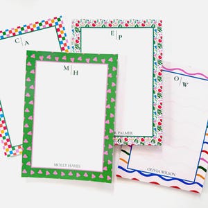 May include: Four personalized notepads with decorative borders. One has a colorful checkerboard pattern, another has a floral design, a third has pink heart shapes, and the last has wavy lines. Each notepad has initials and a name at the bottom.