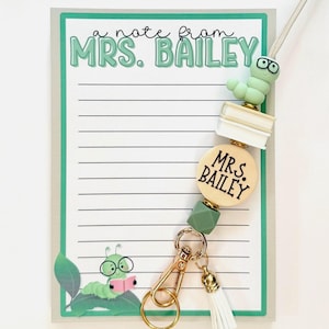 May include: A lanyard with a light green and white beaded design, featuring a worm wearing glasses and books. The lanyard includes a gold clasp, a white tassel, and a wooden bead that says "MRS. BAILEY". A notepad with the text "a note from MRS. BAILEY" is in the background.