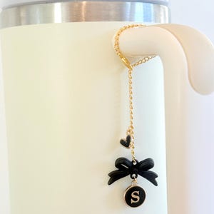 Puede incluir: Un dije de cadena dorada con un lazo negro, un corazón y un colgante circular con la letra "S" cuelga de una taza blanca. El dije está sujeto al asa de la taza. El lazo y el corazón son negros.