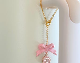 May include: A gold-colored chain with a pink bow charm and a circular pendant with the letter "T" on it. The charm is attached to a white object, possibly a tumbler or water bottle.