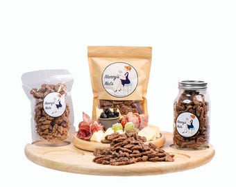 May include: A wooden serving tray with a variety of snacks, including pecan nuts, cheese, and olives. The pecans are in a clear glass jar, a clear plastic bag, and a brown paper bag. The bags and jar have labels that say "Nancy's Nuts".