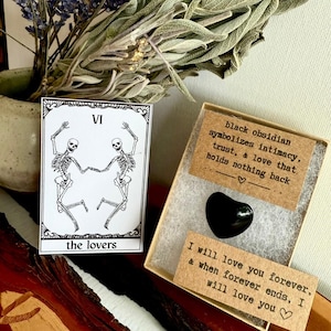 May include: A tarot card with a skeleton couple and the words "the lovers" next to a black heart-shaped obsidian stone in a box. The box contains a card that reads "black obsidian symbolizes intimacy, trust, & love that holds nothing back."