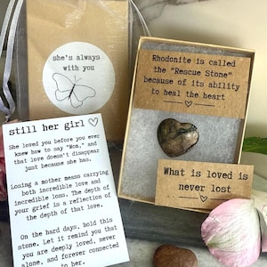 May include: A heart-shaped rhodonite stone inside a gift box with cards featuring text about loss and love. One card reads, Rhodonite is called the Rescue Stone because of its ability to heal the heart. Another card says, still her girl.