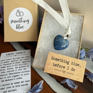 Op de afbeelding: Een hartvormige, blauwe lapis lazuli hanger aan een wit lint, gepresenteerd in een kleine geschenkdoos. De doos bevat een label met de tekst "Something blue, before I do." Een tweede doos met een sticker "something blue" is ook zichtbaar.