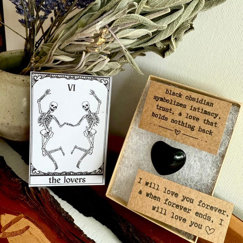 May include: A tarot card with a skeleton couple and the words "the lovers" next to a black heart-shaped obsidian stone in a box. The box contains a card that reads "black obsidian symbolizes intimacy, trust, & love that holds nothing back."
