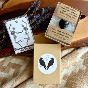 May include: An arrangement featuring a tarot card with skeleton figures labeled "the lovers", a heart-shaped black obsidian stone in a box with text, and a box with a sticker of two ravens. Dried lavender sprigs are also present.