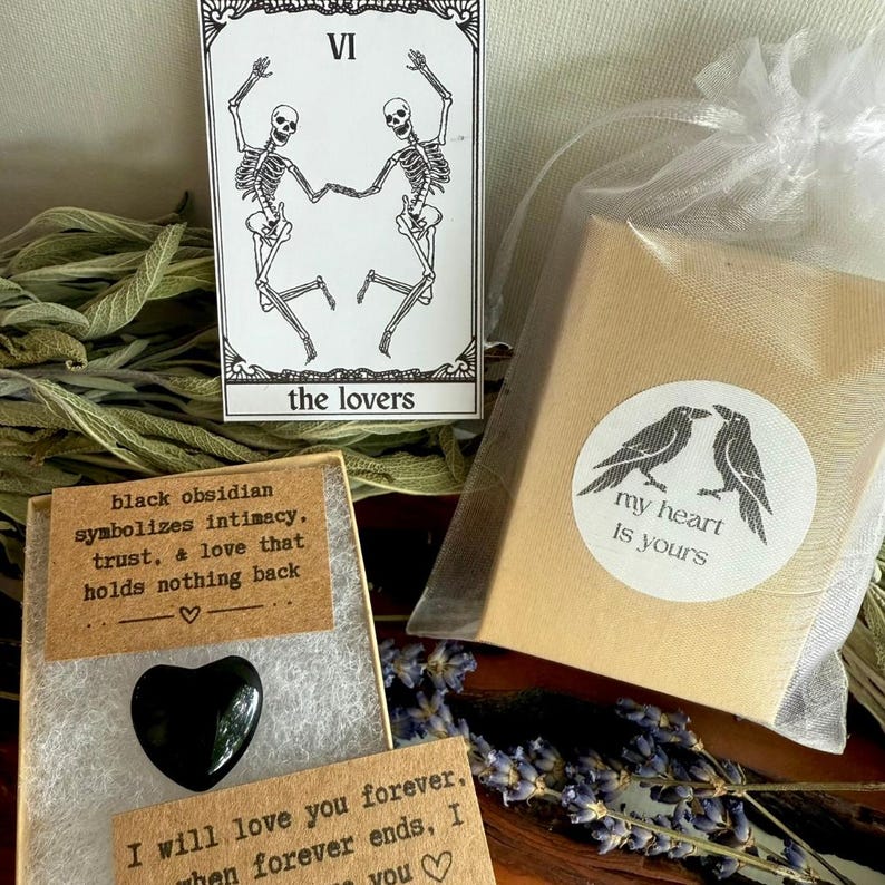 May include: An assortment of items including a tarot card with skeletons, a black heart-shaped obsidian stone, and a small gift box with a sticker that reads "my heart is yours". The items are arranged on a wooden surface.