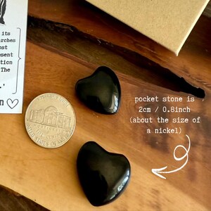 May include: Two polished black heart-shaped pocket stones next to a nickel. The text indicates the stones are 0.8 inches in size, about the size of a nickel. The stones are on a wooden surface.