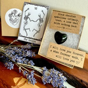 May include: A black heart-shaped obsidian stone in a small box with text that reads "black obsidian symbolizes intimacy, trust, & love that holds nothing back." A tarot card with skeletons dancing and the words "the lovers" is also visible.