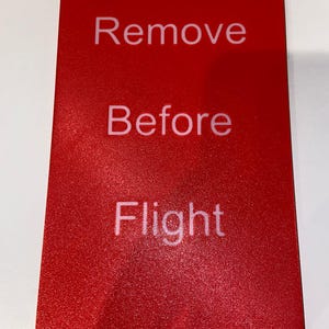 Könnte beinhalten: Ein rotes, rechteckiges Schild mit weißer Aufschrift "Remove Before Flight". Die Oberfläche ist strukturiert. Wird in der Luftfahrt zur Kennzeichnung von vor dem Flug zu entfernenden Gegenständen verwendet.
