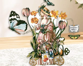 May include: A decorative paper cutout featuring a vintage illustration of a woman in a pink dress surrounded by flowers and butterflies. The cutout includes the text "Jardiniere Royale" and seed packet illustrations. The overall design has a whimsical, garden theme.