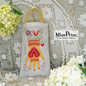 May include: A linen bag with a cross-stitch design featuring a yellow hand holding a red heart with the word "Love". The bag has a yellow handle and the text "Miss Prim. Cross Stitch Pattern".