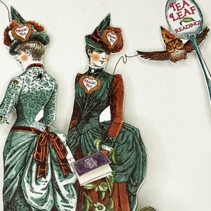 Peut inclure: Illustration vintage de deux femmes en robes et chapeaux, avec l'inscription "Magic Tea" sur leurs poitrines et chapeaux. Un petit hibou tient une cuill&egrave;re avec "Tea Leaf Readings". Un livre avec "Halloween Dance" est &eacute;galement visible.