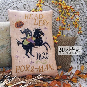 Könnte beinhalten: Ein beigefarbenes Kissen mit einem Kreuzstich-Motiv eines kopflosen Reiters, der ein schwarzes Pferd reitet. Das Design enthält den Text "HEADLESS HORSMAN 1820" und einen Kürbis mit einem Gesicht. Das Kissen liegt auf einer weißen Spitzentischdecke mit einem Zweig gelber Blumen.