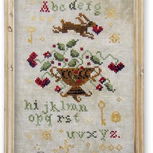 May include: A framed cross-stitch sampler with a white background and colorful embroidery. The sampler features a vase of flowers, a rabbit, and the alphabet. The text "Miss Prim" is embroidered in the bottom right corner.