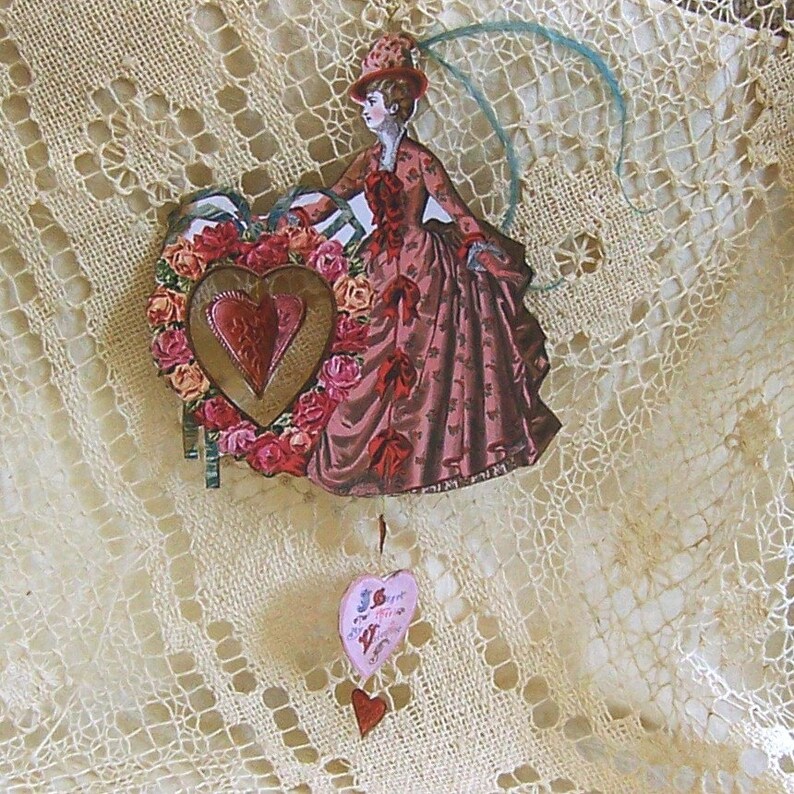 Puede incluir: Una tarjeta vintage de San Valent&iacute;n con una mujer con un vestido rosa y un sombrero sosteniendo una corona de flores en forma de coraz&oacute;n. La tarjeta est&aacute; colgada de un mantel de encaje con una tarjeta m&aacute;s peque&ntilde;a en forma de coraz&oacute;n colgando debajo.
